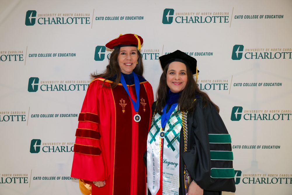 Jennifer Merry and mentor Rebecca Shore pose in front of a UNC Charlotte backdrop in full commencement regalia