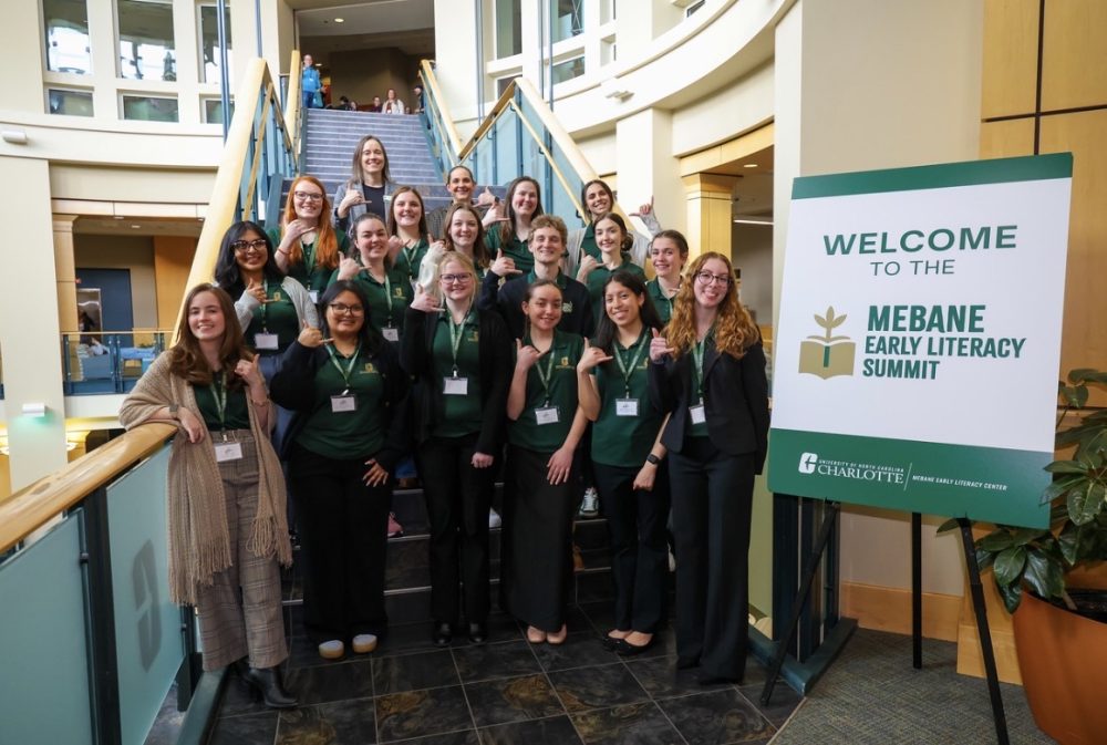 2026 Mebane Scholars pose with their hands forming pickaxes on a staircase with mentors Miranda Fitzgerald and Jennifer Merry next to a sign for the Mebane Early Literacy Summit