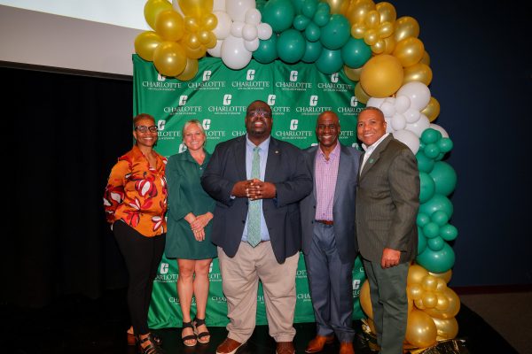 Alumnus Clyde Wilson poses with his father Clyde Wilson Sr., mentor Lisa Merriweather, director Elisabeth Wilson, and Dean Butler in front of a green background and Charlotte-themed balloon arch, celebrating his award